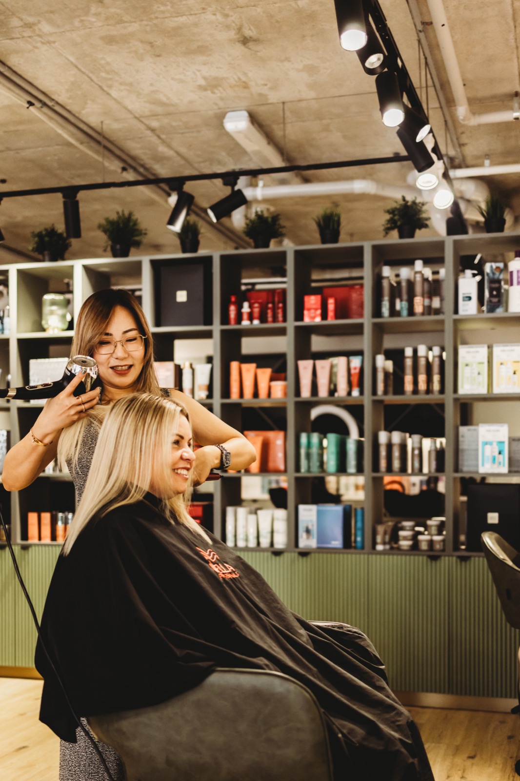 Hair Avenue stylist blow-drying a smiling client in the chair with the product wall behind